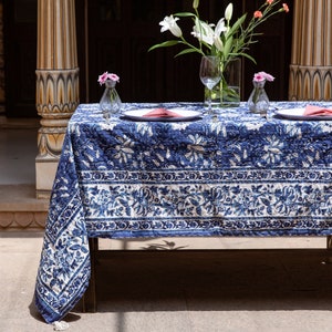 Puede incluir: Un mantel de mesa con estampado azul y blanco con un dise&ntilde;o floral, colocado sobre una mesa con dos cubiertos, cada uno con un plato blanco, una servilleta rosa y un vaso de agua. El mantel tiene un borde con flecos.
