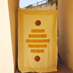 Puede incluir: Una manta de algod&oacute;n amarilla con un patr&oacute;n geom&eacute;trico en un tono de amarillo m&aacute;s oscuro. El patr&oacute;n presenta un c&iacute;rculo, cuatro rect&aacute;ngulos horizontales y un segundo c&iacute;rculo en la parte inferior.