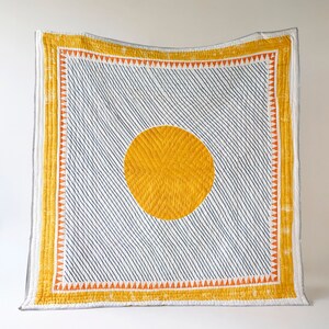 May include: A handmade quilt featuring a large yellow sun design on a white background with diagonal navy blue lines. The quilt has a yellow border with orange triangles and is a decorative home textile.