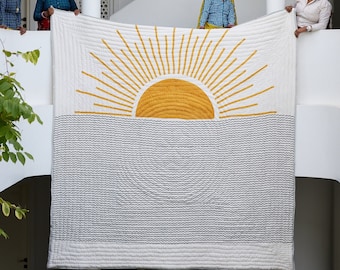May include: Four women are holding up a white and yellow quilt with a sun and waves design. The quilt is hanging from a white railing.