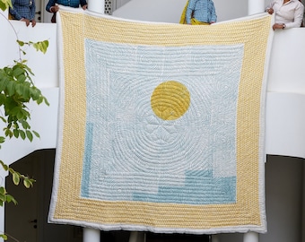 May include: Four women stand on a white balcony holding up a large, rectangular quilt. The quilt is white with a blue and yellow design. The design features a large yellow circle in the center and a repeating pattern of swirls.
