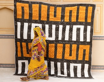 May include: A woman in a yellow and floral sari stands in front of a large black and orange quilt. The quilt has a geometric pattern of squares and rectangles.