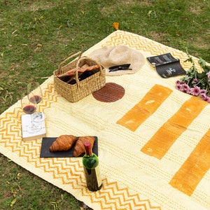 Puede incluir: Una manta de picnic amarilla y naranja con un borde de patr&oacute;n de chevron. La manta est&aacute; extendida sobre hierba verde y tiene una cesta de mimbre, una botella de vino, vasos de vino, croissants, un libro y gafas de sol encima.
