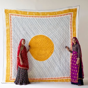 May include: Large, handmade quilt with a white background and diagonal navy blue stripes. A large yellow circle is centered on the quilt. The quilt has a yellow border with orange triangles. Two people stand on either side of the quilt.