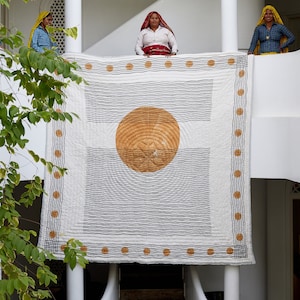 Pode incluir: Uma colcha listrada branca e preta com um grande desenho de sol amarelo redondo no centro. A colcha está pendurada em uma balaustrada branca e tem uma borda de pequenos círculos amarelos.