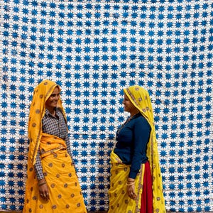 May include: Two women in traditional Indian attire stand before a large blue and white patterned textile. One woman wears a yellow sari with a matching head covering, while the other wears a blue top and a yellow sari with a red underskirt. The textile has a repeating star pattern.