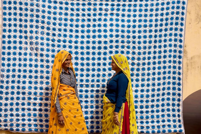 May include: Two women in traditional Indian attire stand before a large blue and white patterned textile. One woman wears a yellow sari with a matching head covering, while the other wears a blue top and a yellow sari with a red underskirt. The textile has a repeating star pattern.