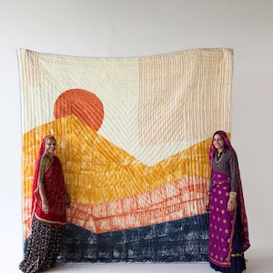 May include: A large, rectangular quilt with a landscape design featuring a vibrant orange sun, yellow and orange mountains, and a dark blue base. The quilt is displayed against a white background, with two people standing on either side.