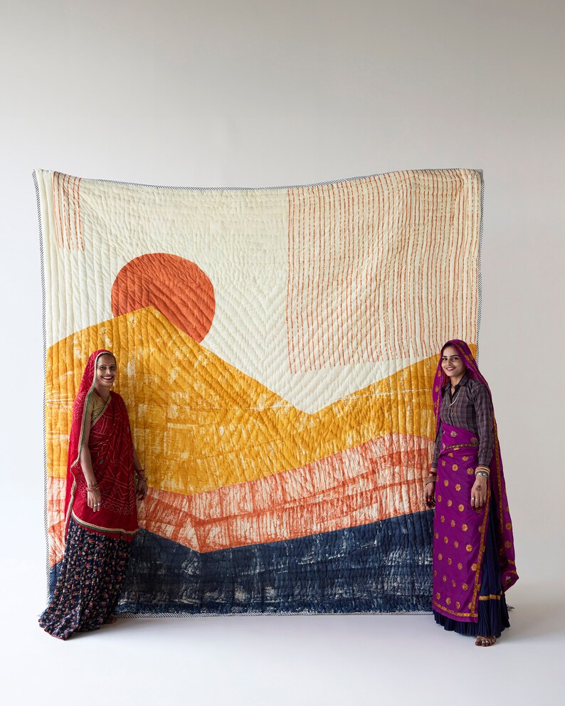 May include: A large, rectangular quilt with a landscape design featuring a vibrant orange sun, yellow and orange mountains, and a dark blue base. The quilt is displayed against a white background, with two people standing on either side.