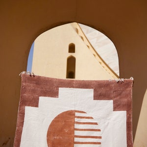 May include: A white and brown woven rug with a geometric pattern of circles and squares. The rug is hanging in a doorway with a building visible in the background.