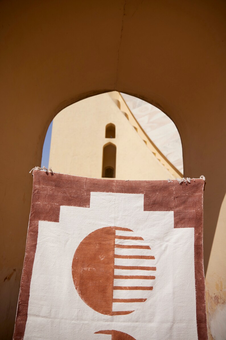 May include: A white and brown woven rug with a geometric pattern of circles and squares. The rug is hanging in a doorway with a building visible in the background.