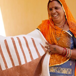 May include: A woman wearing a bright orange headscarf holds a white and brown striped rug with a brown semi-circle design. She is wearing a gold necklace and bracelets.