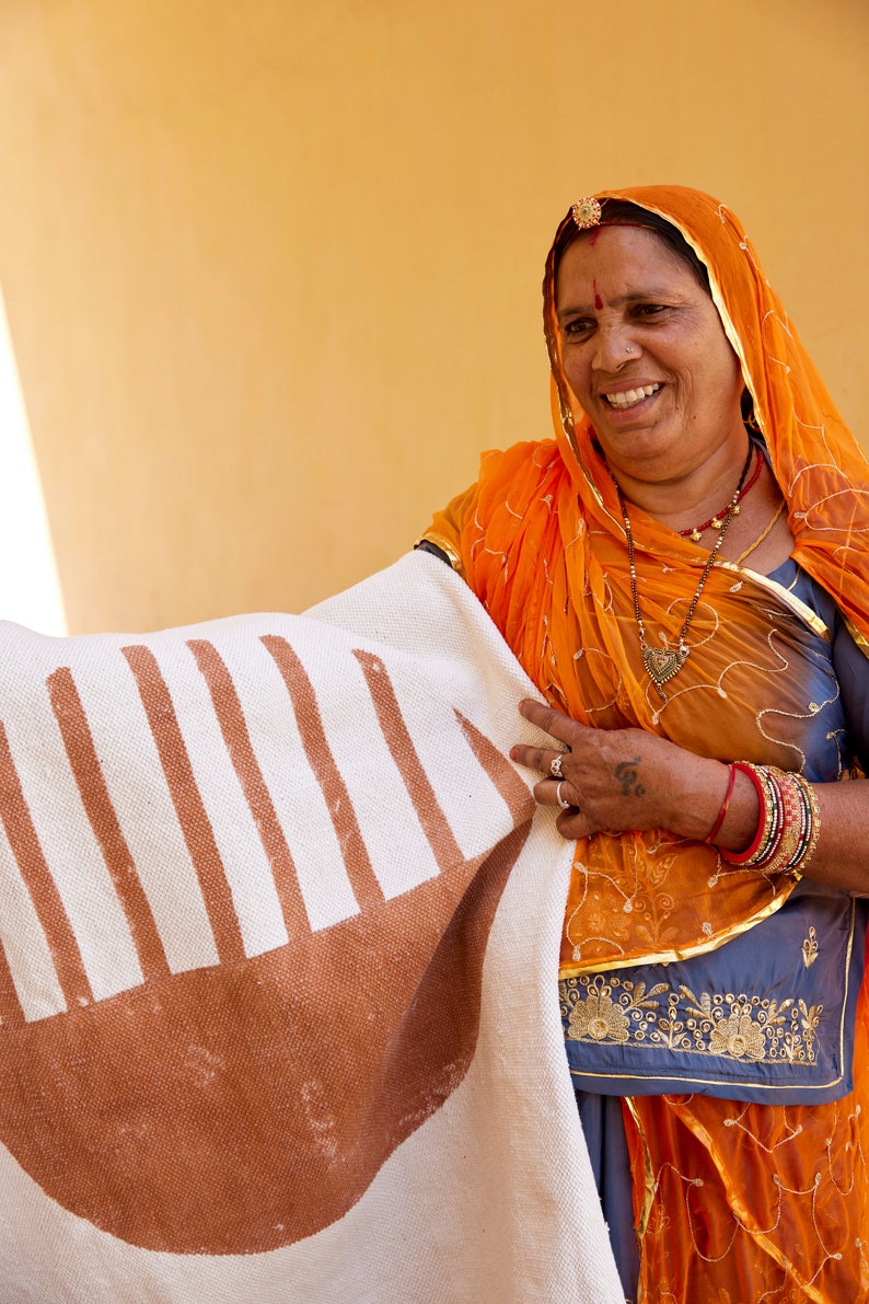 May include: A woman wearing a bright orange headscarf holds a white and brown striped rug with a brown semi-circle design. She is wearing a gold necklace and bracelets.
