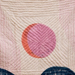 May include: A cream-colored quilt with a pink and orange sun design and blue mountain shapes at the bottom. The quilt has a quilted pattern with straight and diagonal lines. The sun is a circle with a cross-stitch pattern.