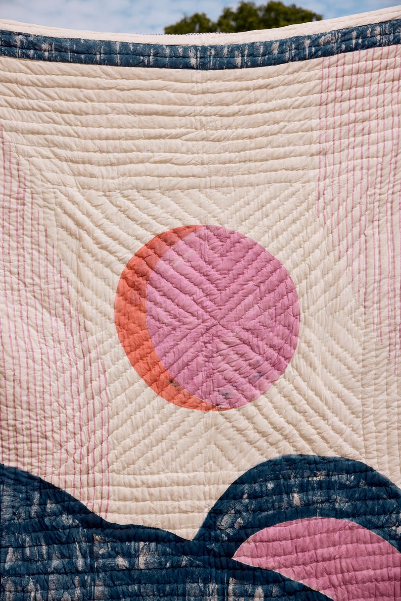 May include: A cream-colored quilt with a pink and orange sun design and blue mountain shapes at the bottom. The quilt has a quilted pattern with straight and diagonal lines. The sun is a circle with a cross-stitch pattern.