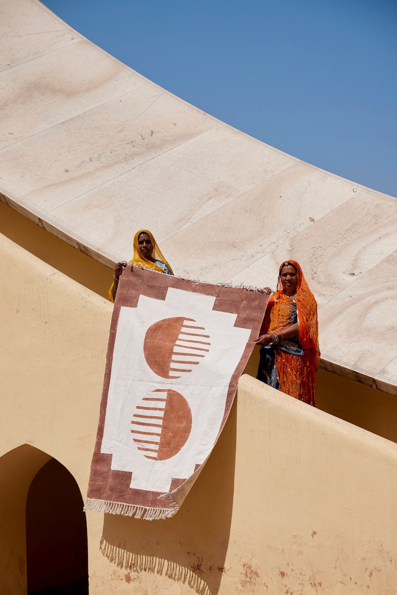 May include: Two women in colorful saris stand on a beige wall holding a large, rectangular rug with a brown and white geometric pattern. The rug has two large, half-circle shapes with a striped pattern.