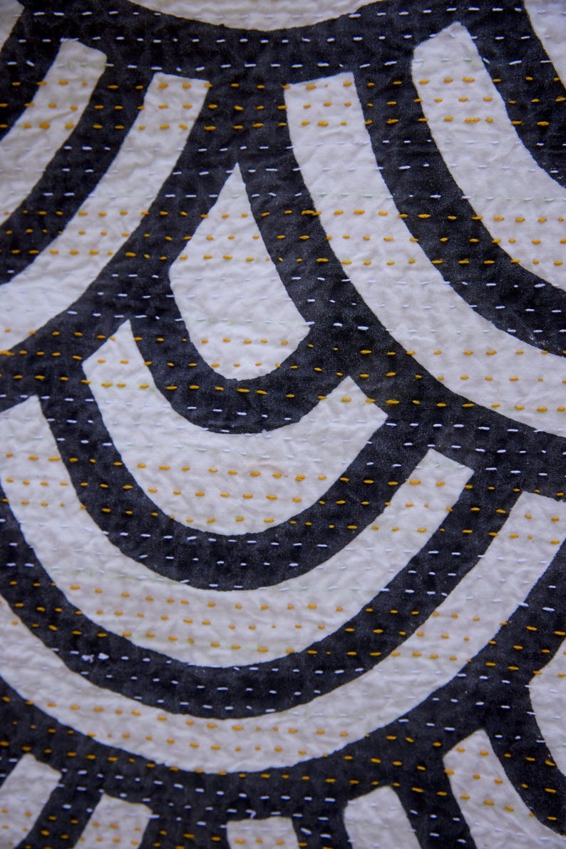 Peut inclure: Un textile pr&eacute;sentant un motif r&eacute;p&eacute;titif de formes courbes noires sur un fond blanc. Le tissu est orn&eacute; de rang&eacute;es de petits points de couture horizontaux en fil blanc et jaune moutarde, cr&eacute;ant un aspect textur&eacute; et matelass&eacute; sur la surface.