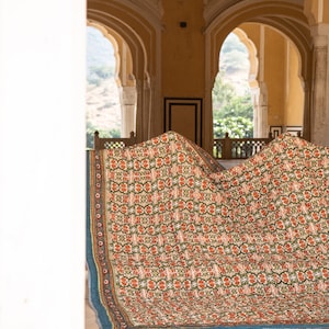 Pu&ograve; includere: Una trapunta con motivi verdi, arancioni e bianchi &egrave; piegata e adagiata su un pavimento in un edificio con porte ad arco. La trapunta ha un bordo blu.