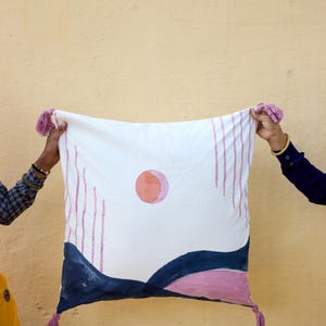 May include: White square pillow with a hand-painted design featuring a pink sun, blue and pink landscape, and vertical pink lines. The pillow has pink tassels on each corner.