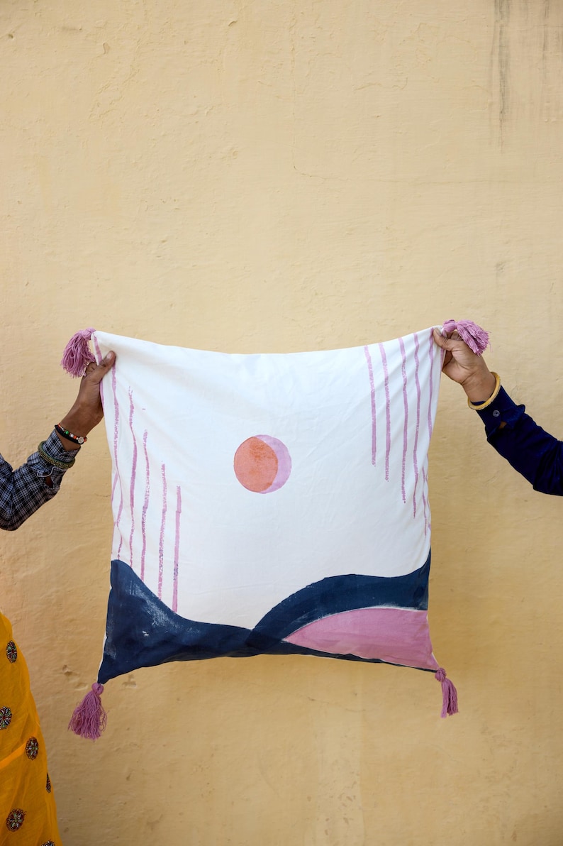 May include: White square pillow with a hand-painted design featuring a pink sun, blue and pink landscape, and vertical pink lines. The pillow has pink tassels on each corner.