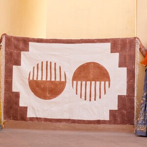 May include: Two women in traditional Indian clothing hold up a white fabric rug with a brown border and two brown circles with vertical lines. The rug has a geometric pattern.