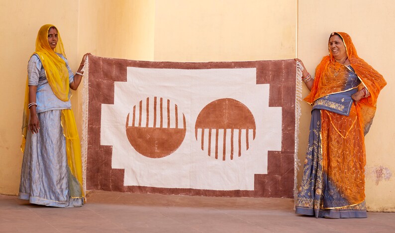 May include: Two women in traditional Indian clothing hold up a white fabric rug with a brown border and two brown circles with vertical lines. The rug has a geometric pattern.