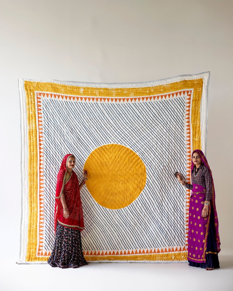 May include: Large, handmade quilt with a white background and diagonal navy blue stripes. A large yellow circle is centered on the quilt. The quilt has a yellow border with orange triangles. Two people stand on either side of the quilt.