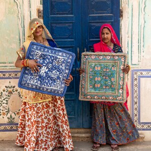 May include: Two women stand in front of a blue door, each holding a colorful patterned cushion. The cushions have a floral design and are framed with a decorative border. The women are wearing traditional Indian clothing.