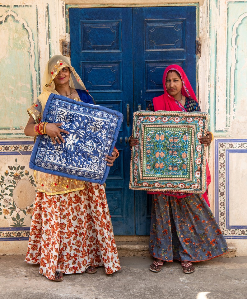 May include: Two women stand in front of a blue door, each holding a colorful patterned cushion. The cushions have a floral design and are framed with a decorative border. The women are wearing traditional Indian clothing.