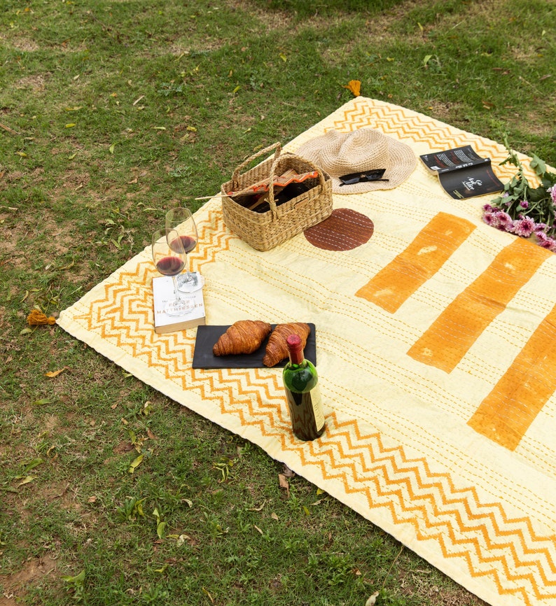 Puede incluir: Una manta de picnic amarilla y naranja con un borde de patr&oacute;n de chevron. La manta est&aacute; extendida sobre hierba verde y tiene una cesta de mimbre, una botella de vino, vasos de vino, croissants, un libro y gafas de sol encima.