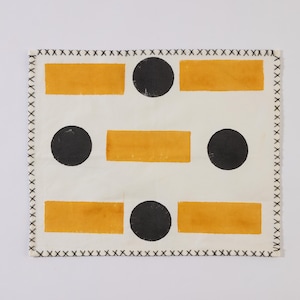 Puede incluir: Textil rectangular con fondo blanco, con un patrón de círculos negros y rectángulos naranjas. Los bordes están acabados con un borde cosido negro. El diseño es simple y geométrico.