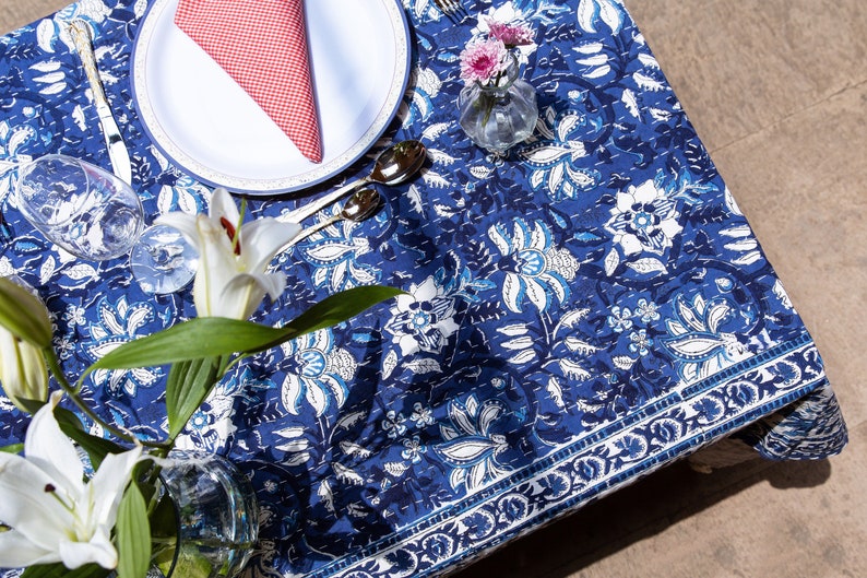 Puede incluir: Un mantel de mesa floral azul y blanco con una servilleta a cuadros rojos y blancos sobre un plato blanco. Hay lirios blancos en un jarr&oacute;n transparente sobre la mesa.