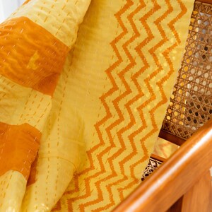 Puede incluir: Una colcha kantha amarilla y naranja con un patr&oacute;n de chevron. La colcha est&aacute; doblada y drapeada sobre una silla de madera con un asiento de ca&ntilde;a tejido.