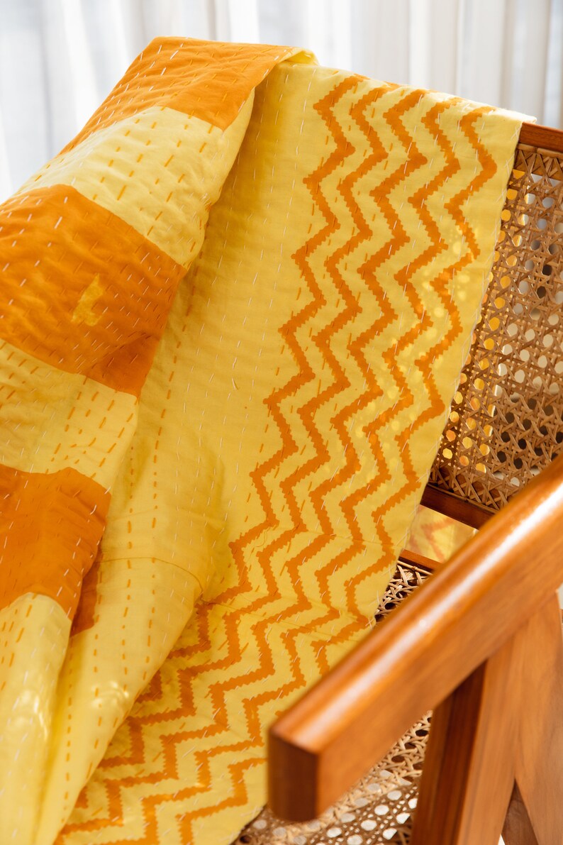 Puede incluir: Una colcha kantha amarilla y naranja con un patr&oacute;n de chevron. La colcha est&aacute; doblada y drapeada sobre una silla de madera con un asiento de ca&ntilde;a tejido.