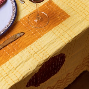 Puede incluir: Un mantel amarillo con un patr&oacute;n geom&eacute;trico marr&oacute;n y naranja. Un vaso de vino tinto se encuentra sobre el mantel, junto a un plato blanco con una servilleta roja.