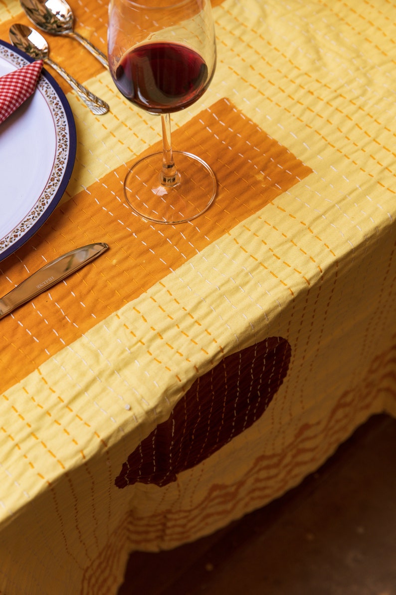 Puede incluir: Un mantel amarillo con un patr&oacute;n geom&eacute;trico marr&oacute;n y naranja. Un vaso de vino tinto se encuentra sobre el mantel, junto a un plato blanco con una servilleta roja.