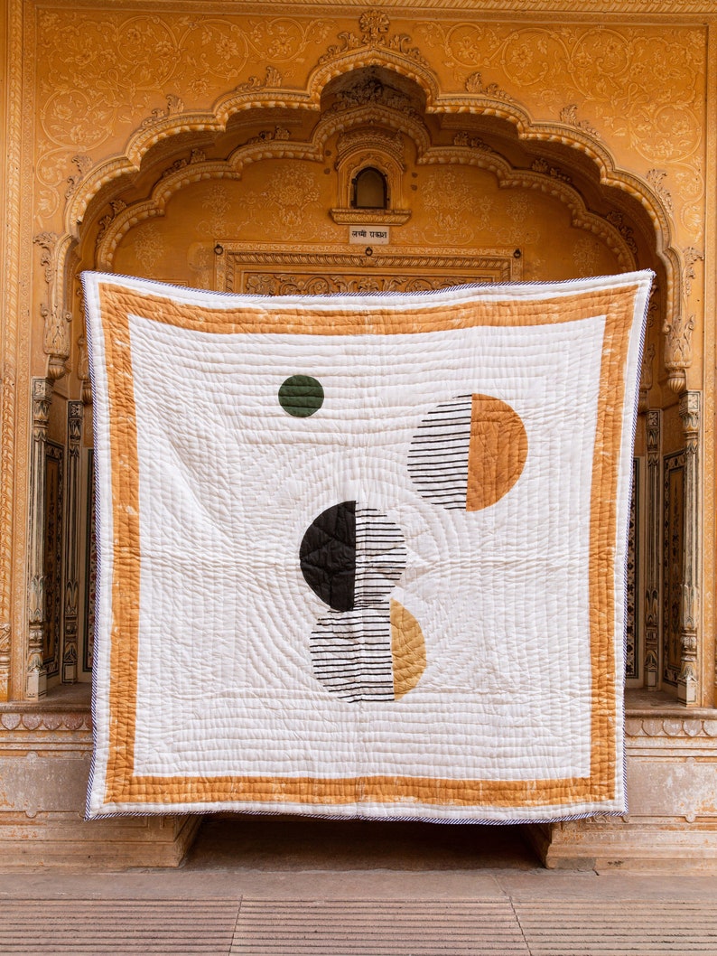 May include: A white quilt with a geometric design featuring black, white, yellow, and green circles. The quilt is hanging on a wall with a yellow background.