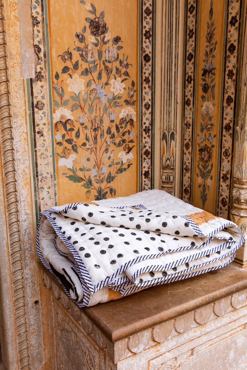 May include: A white and black polka dot quilt with a blue and white striped border is folded and placed on a stone bench. The bench is in front of a wall with intricate floral designs painted in yellow, green, and brown.