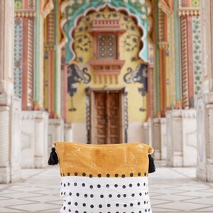 May include: A mustard yellow and white throw pillow with black polka dots and black tassels. The pillow is in front of a colorful, ornate doorway.