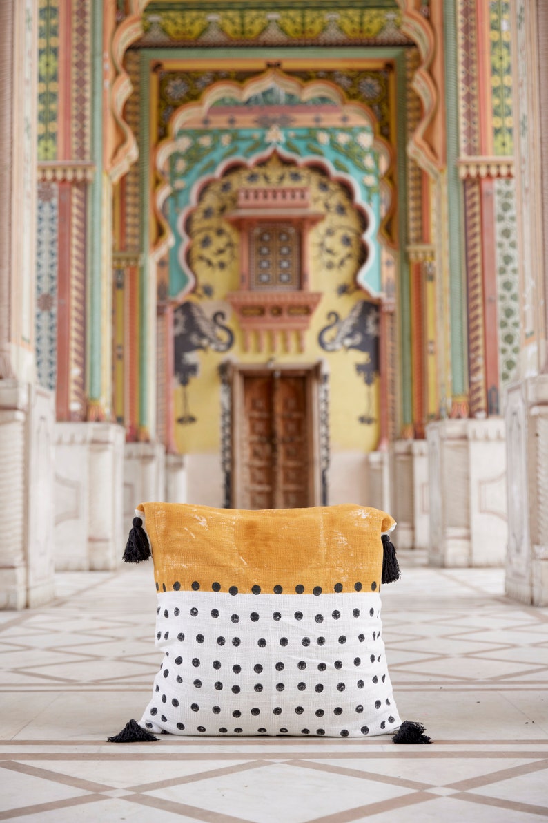 May include: A mustard yellow and white throw pillow with black polka dots and black tassels. The pillow is in front of a colorful, ornate doorway.