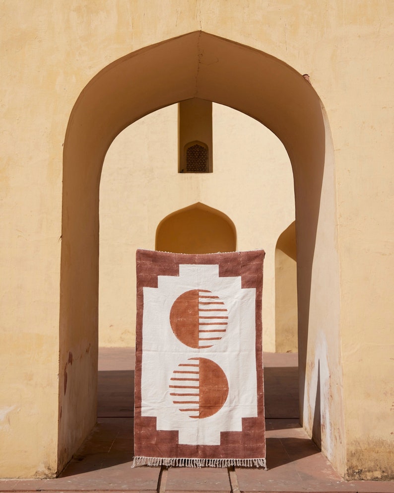May include: A white and brown geometric rug with a repeating pattern of half circles. The rug is hanging in a doorway with a yellow archway.