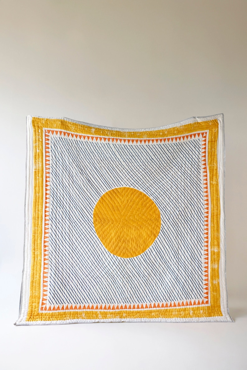 May include: A handmade quilt featuring a large yellow sun design on a white background with diagonal navy blue lines. The quilt has a yellow border with orange triangles and is a decorative home textile.