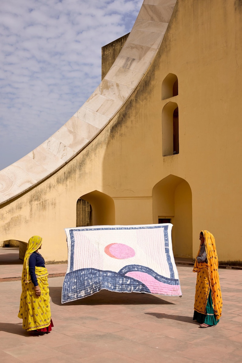 May include: A rectangular quilt with a landscape design featuring a pink sun, blue waves, and white background is held up by two people. The quilt is displayed in front of a yellow building with arched windows and a large, curved structure. The people are wearing traditional Indian clothing.