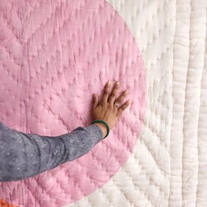 May include: A quilted textile featuring a large pink circle design on a white background. The quilt has a stitched pattern and a hand is resting on the pink circle. The quilt is a soft, textured fabric.