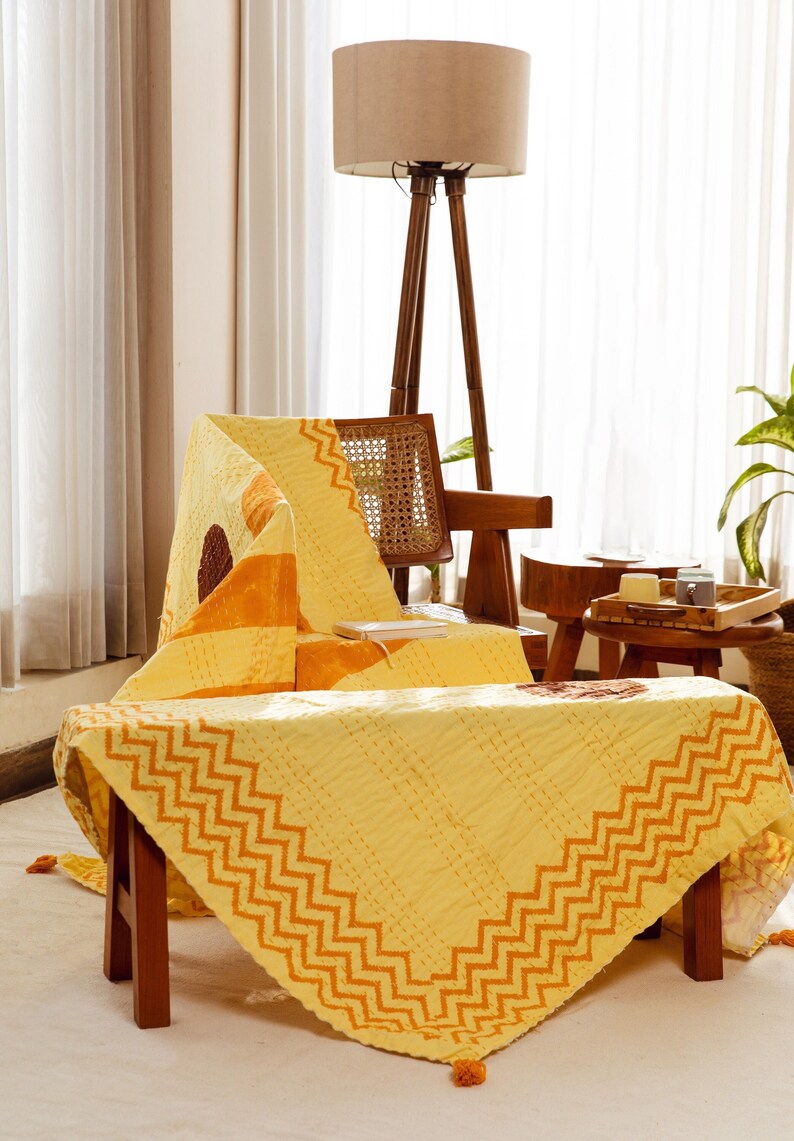 Puede incluir: Una colcha kantha amarilla y naranja con un patr&oacute;n en zigzag sobre un taburete de madera. La colcha est&aacute; doblada y drapeada sobre una silla en el fondo.