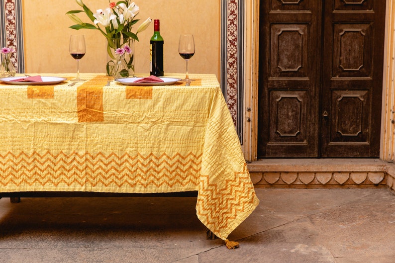 Puede incluir: Un mantel amarillo con un patr&oacute;n geom&eacute;trico en naranja, preparado para una cena rom&aacute;ntica con dos copas de vino, una botella de vino y platos. El mantel tiene un borla en el extremo.