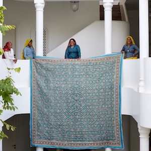 Puede incluir: Cuatro mujeres se paran en un balc&oacute;n blanco sosteniendo una gran colcha estampada en azul y verde. La colcha tiene un dise&ntilde;o floral y un borde blanco.