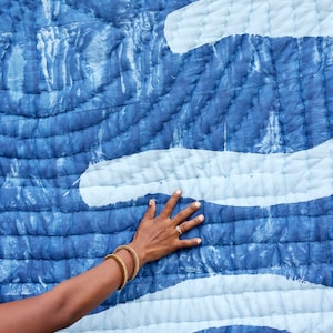 K&ouml;nnte beinhalten: Blau-wei&szlig; gesteppter Textil mit wellenartigem Muster. Die Steppdecke hat einen dunkelblauen Hintergrund mit helleren blauen und wei&szlig;en Akzenten. Die Textur ist sichtbar, mit gen&auml;hten Linien, die einen gesteppten Effekt erzeugen.