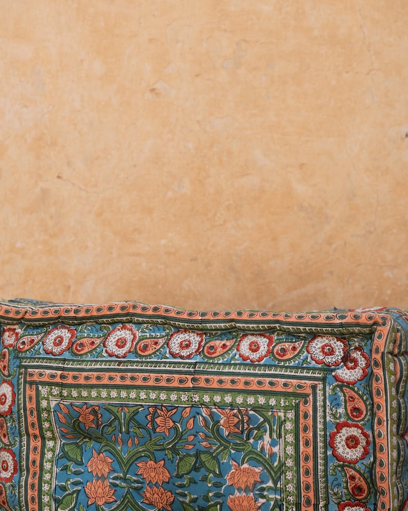 May include: A close-up of a colorful patterned cushion with a floral design. The cushion is made of fabric and has a light blue background with orange, green, and white accents.