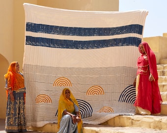 May include: Three women stand in front of a large white and blue patterned quilt. The quilt has a striped design with a wavy pattern. The women are wearing colourful clothing.